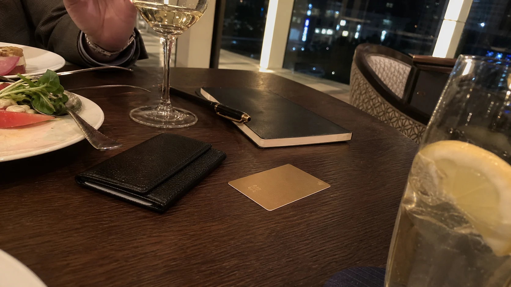 夜景の見えるホテルダイニングで食事、グラス、旅行ノート、無地のゴールドカード風プレートが置かれた特典利用イメージ